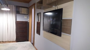 Een slaapkamer met een bed, een aan de muur bevestigde televisie, houten kasten en een raam met gordijnen.