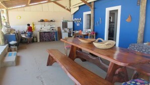 A rustic kitchen with wooden benches and a table, a person cooking, and a blue wall with a door. - Paraiso in Bahia - house by the sea in the middle of coconut trees (Belmonte)