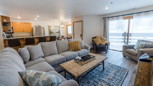 Un salon moderne avec un canapé gris, une table basse et un coin repas doté de meubles de rangement en bois et d’un réfrigérateur.