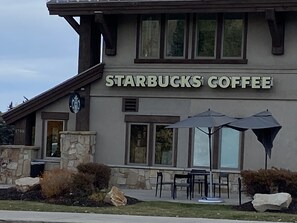 Un bâtiment de Starbucks avec une façade en pierre, de grandes fenêtres, des places assises extérieures avec des parasols, et un pilier en pierre portant le numéro 1700.