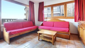 Une pièce chaleureuse avec un canapé en bois et une petite table en bois, un balcon avec des rideaux rouges et une vue sur un immeuble doté d’un balcon.