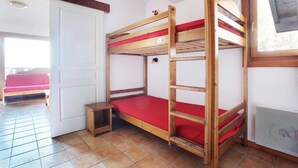 Un lit superposé avec un matelas rouge, une petite table de chevet en bois et une fenêtre donnant sur les arbres.