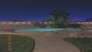 Vista noturna de uma piscina com uma espreguiçadeira vermelha e palmeiras.