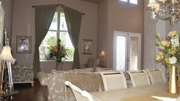 Uma sala de jantar com mesa posta para quatro pessoas, rodeada por cadeiras estofadas. Há um candelabro sobre a mesa e uma janela com cortinas à esquerda.