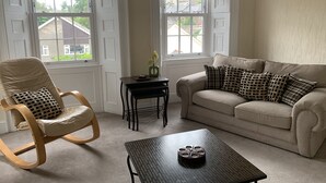 Un soggiorno con un divano color beige, una poltrona a dondolo in legno, un tavolino e una finestra che si affaccia su case.