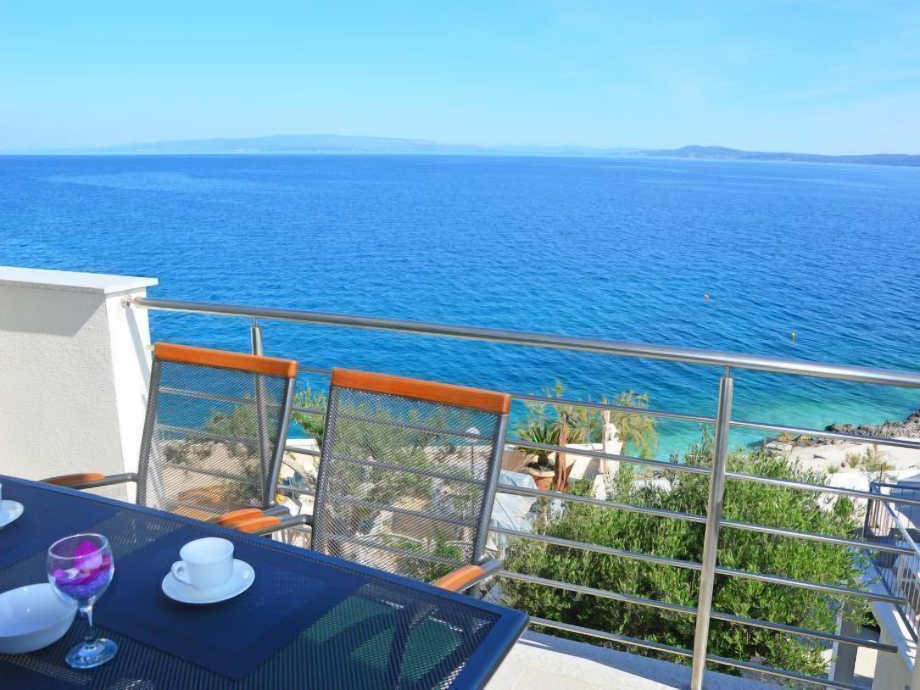 A balcony with a table set for two, overlooking a clear blue sea and distant land.