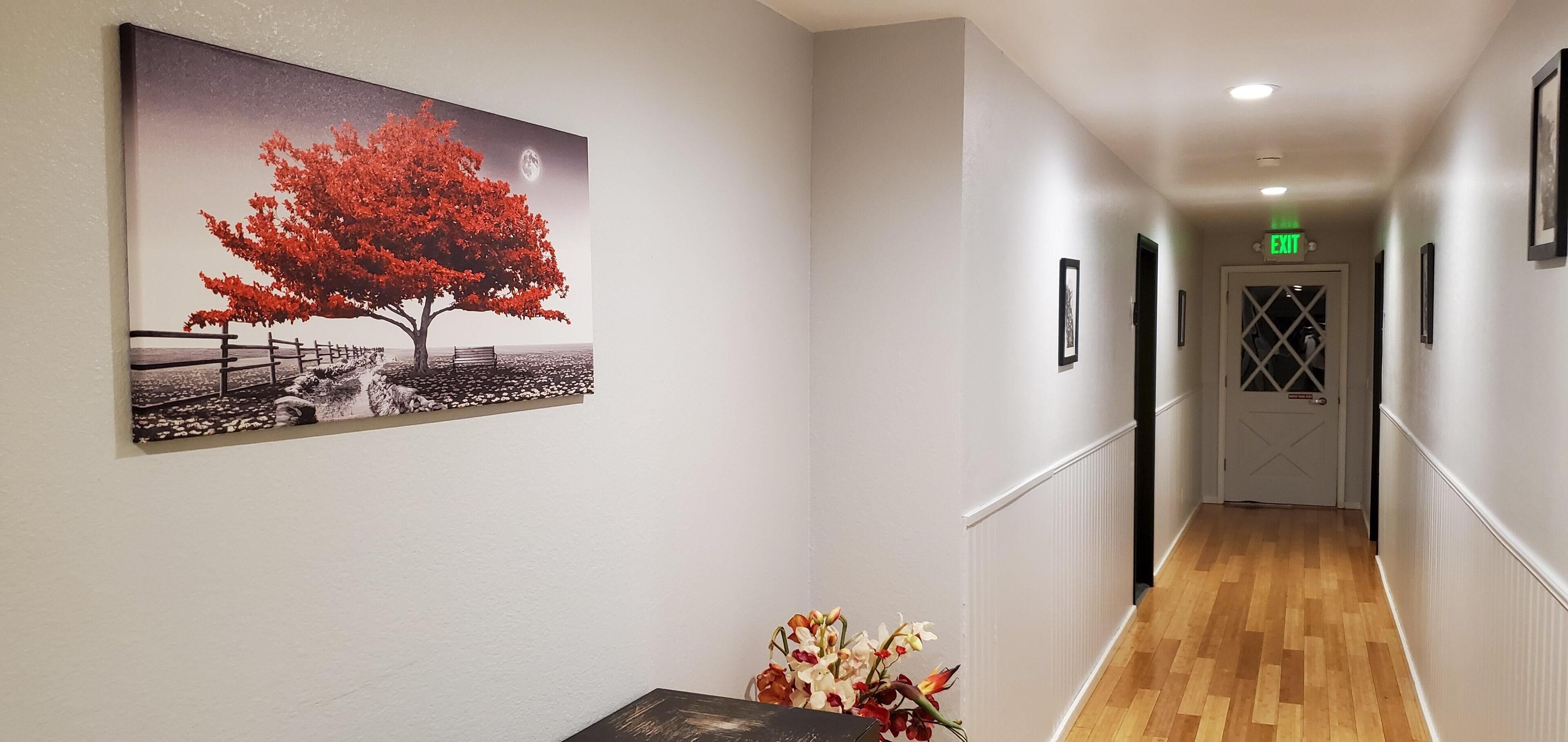 A hallway with a wooden floor, a bench, a framed picture of a tree, and a vase of flowers.