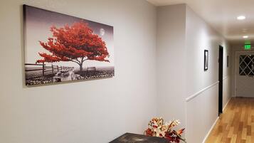 A hallway with a wooden floor, a bench, a framed picture of a tree, and a vase of flowers.