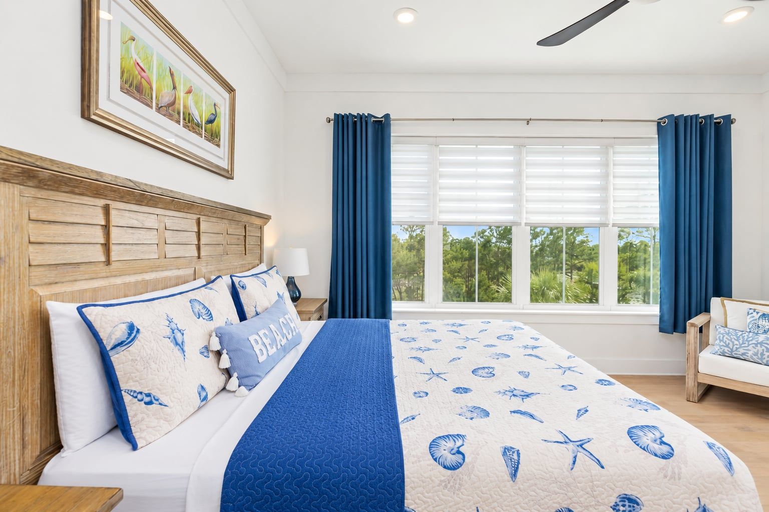 A bedroom with a large bed, a headboard, a nightstand, a lamp, a window with curtains, and a view of greenery outside.