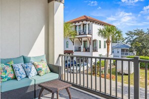 A balcony with a wicker sofa and a small table, overlooking a street with parked cars and a building with a red roof. - Sandy Bottoms - 4BR/3.5BA Starview Terrace Home - Sleeps 13 - Golf Cart & Community Pool (Santa Rosa Beach)