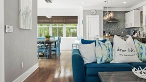 A modern living room with a blue sofa, dining area with blue chairs, and a kitchen with white cabinets and a backsplash.
