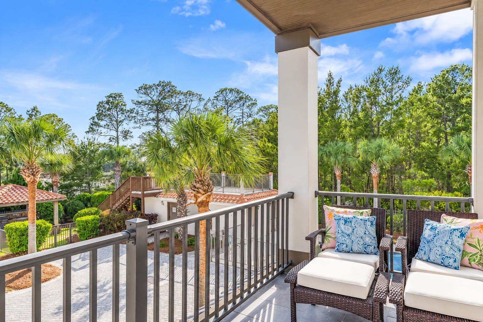 A balcony with wicker furniture and a view of a landscaped garden with trees and a building in the background.