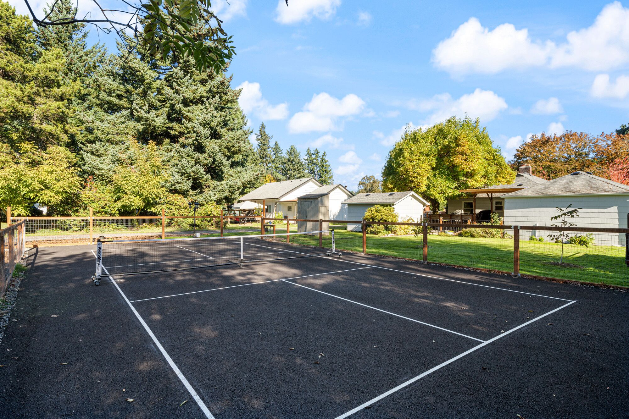 A tennis court surrounded by a fence, with a row of houses and trees in the background.