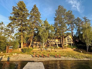 A lakeside cabin with a dock, surrounded by tall trees and a clear sky. - Peaceful Island Retreat on Foys Lake MT – Hot Tub, Private Dock, Beach & Kayaks! (Kalispell)