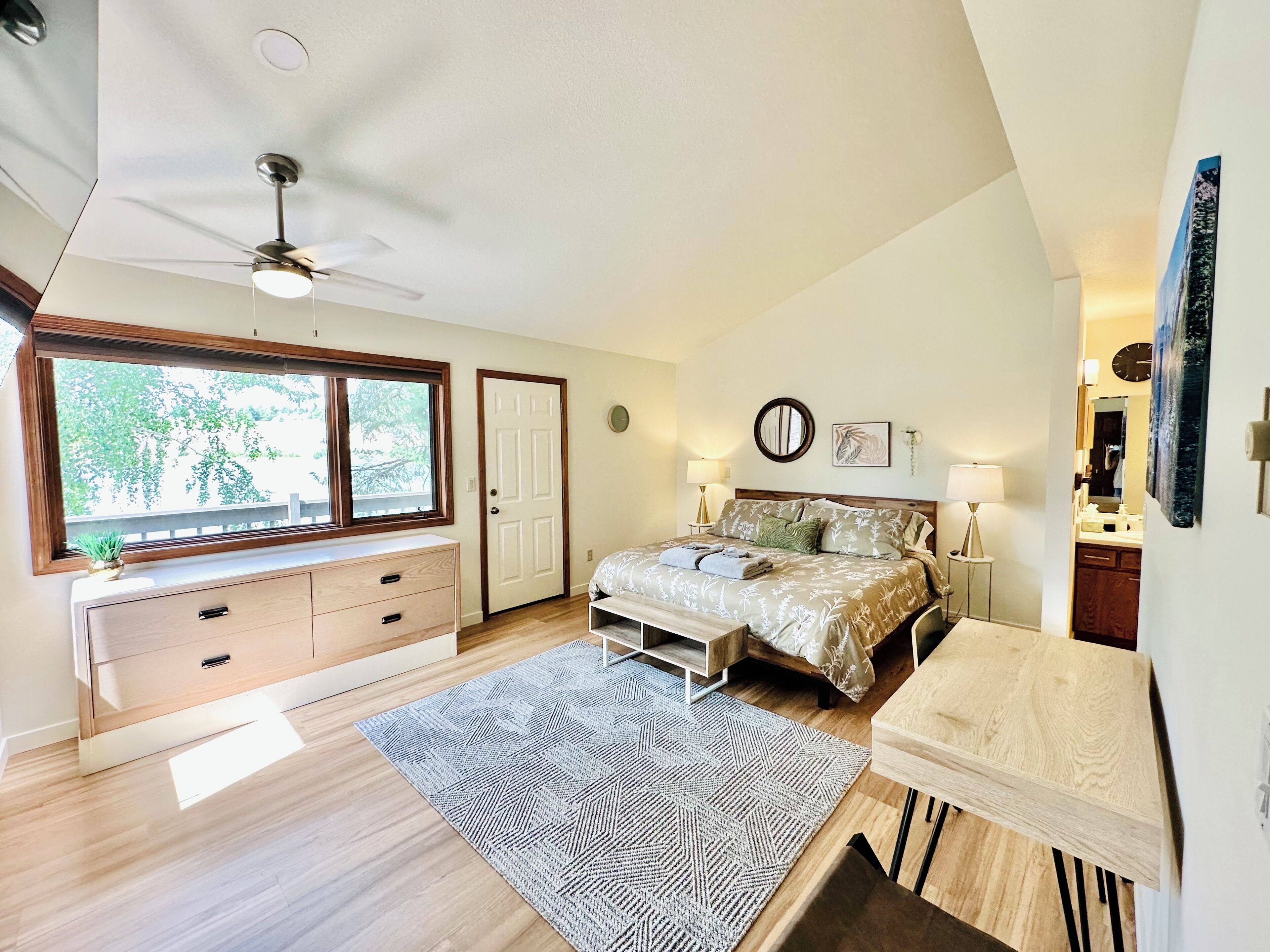 A bedroom with a bed, bedside tables, a desk, a ceiling fan, a window, and a door.