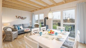 Ein gemütliches Wohnzimmer mit einer Couch, einem Korbsessel, einem Esstisch mit Schüsseln und Tassen und einem großen Fenster mit Blick auf Grünflächen.