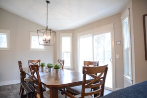A dining area with a wooden table and chairs, a hanging light fixture, and large windows providing natural light.
