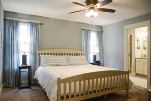 A bedroom with a wooden bed, two bedside tables with lamps, a ceiling fan, and a view of a bathroom through an open door.