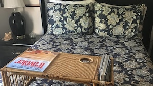 Ein Bett mit dunkler Blumenbettdecke, ein gewebter Nachttisch mit einer Zeitschrift und eine Lampe auf einem Beistelltisch.
