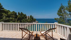 Un balcon avec deux chaises en bois et une table, d’où l’on peut admirer une mer d’un bleu éclatant et une côte rocheuse bordée d’arbres.