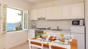 Une cuisine compacte avec des meubles blancs, une table à manger dressée pour le petit-déjeuner et une vue sur la mer par la fenêtre.
