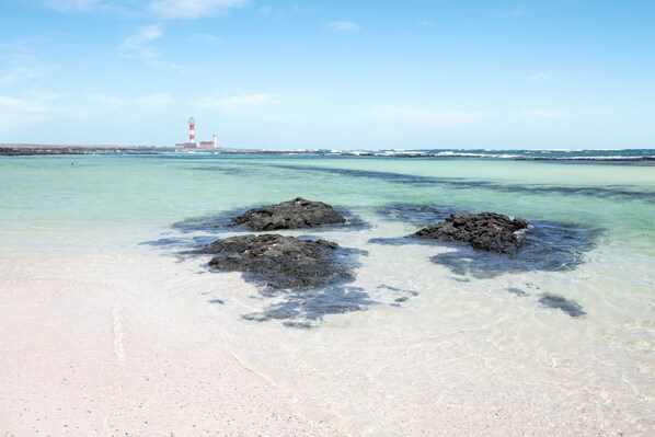 A beach with clear water, three large rocks, and a lighthouse in the distance.