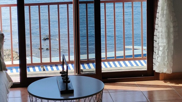 A room with a view of the ocean, a balcony with a railing, and a table with a remote control.