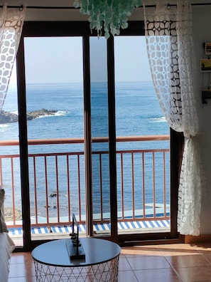 A room with a view of the ocean, a balcony with a railing, and a table with a remote control.