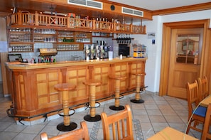 Zona de bar con taburetes de madera, estantes con botellas, vasos y una caja registradora.