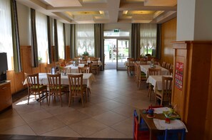 Un comedor con varias mesas y sillas, un televisor a la izquierda y vistas al exterior a través de amplias ventanas.