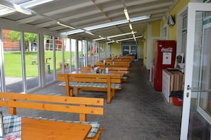 Un área de estar al aire libre techada con bancos y mesas de madera, un refrigerador de Coca-Cola y vistas a un espacio cubierto de césped con un parque infantil.