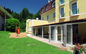 Un bâtiment jaune avec un toit rouge, une terrasse couverte et un espace de jeux pour enfants.