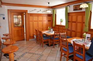 A dining area with wooden furniture, a table with chairs, a bar area with stools, and a window with curtains. - Standard Single Room - Kirchenwirt Aigen (Aigen im Ennstal)