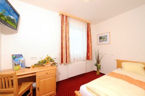 A hotel room with a bed, a desk with a chair, a radiator, a window with curtains, a framed picture on the wall, and a vase with flowers. - Comfort double room - Kirchenwirt Aigen (Aigen im Ennstal)