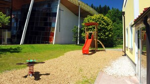 Un parque infantil con tobogán y columpios, un edificio con ventanas amplias y un cielo despejado.