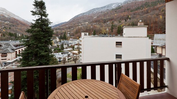 Un balcon avec une table et des chaises en bois, donnant sur une ville avec ses bâtiments et une montagne en arrière-plan.
