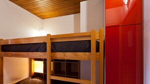 Un lit superposé avec une structure en bois et un matelas, une armoire rouge et une fenêtre laissant entrer la lumière.