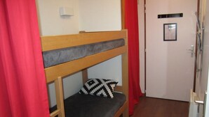 Una cama litera con un cojín estampado, una puerta blanca con un letrero y cortinas rojas.