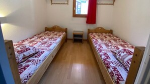Et lille, rent værelse med to træsenge, et lille natbord, et vindue med rødt gardin og træhylder over sengene.