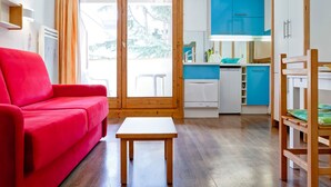 A compact living space with a red sofa, a small wooden table, a kitchenette with blue cabinets, and a sliding glass door leading to an outdoor area. - 2* Studio with Wifi in Brides-les-Bains Center (Brides-les-Bains)