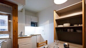 A compact kitchen with a sink, microwave, and wooden cabinets. A dining table with chairs is set for two, and a shelf with dishes is visible. - 2* Studio with balcony, parking and wifi in the heart of Brides-les-Bains (Brides-les-Bains)