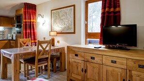 Un cuarto acogedor con una mesa de madera para comer, sillas, un televisor sobre un mueble de madera, una ventana con cortinas y una zona de cocina al fondo.