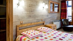 Un dormitorio con cabecera de madera, cama con colcha colorida, lámparas de noche, un espejo, una silla y una ventana con cortinas.