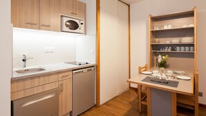 A compact kitchen with wooden cabinets, a built-in microwave, a sink, and a dishwasher. There is a dining area with a table set for two, and a shelf with dishes and glasses. - Studio for 4 with Balcony, WiFi & Parking (Brides-les-Bains)