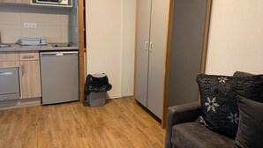 A compact living space with a kitchenette, a grey wardrobe, a small trash bin, and a grey sofa with patterned cushions. - Studio for 4 with Balcony, WiFi & Parking (Brides-les-Bains)