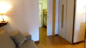 A room with a wooden floor, a grey sofa, a bedside table with a lamp, and a door leading to a bedroom with a bunk bed. - Studio for 4 with Wifi and Parking - Brides-les-Bains (Brides-les-Bains)