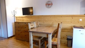 A wooden dining table with chairs, a wall clock, a television, and a trash bin in a room with wooden paneling. - Studio for 4 with Wifi and Parking - Brides-les-Bains (Brides-les-Bains)