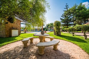 Um jardim com uma fonte de pedra, vegetação exuberante e uma área de piscina ao fundo.