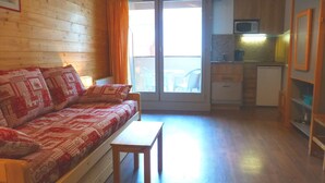 A cozy living room with a wooden sofa, a small table, and a kitchen area with cabinets and a microwave. - Studio for 4 - City Center, Balcony, Wi-Fi (Brides-les-Bains)