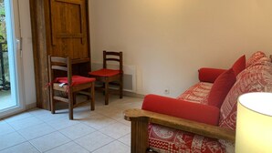 Ein Zimmer mit einer Holztür, zwei Holzsesseln mit roten Kissen, einem roten, gemusterten Sofa und einer Lampe.